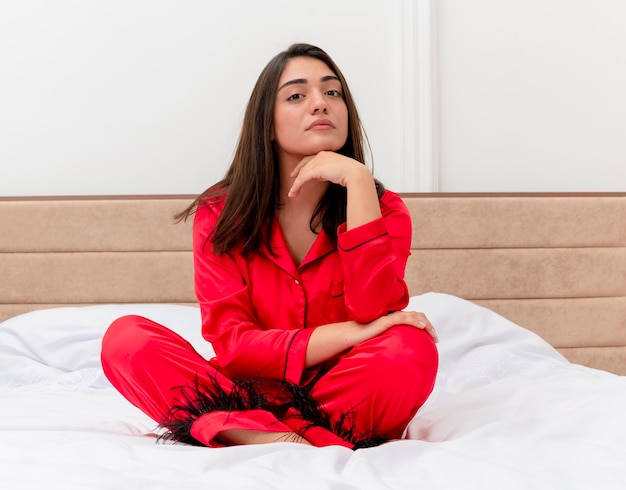 Giovane bella donna in pigiama rosso che si siede sul letto che guarda l'obbiettivo con la faccia seria nell'interno della camera da letto su sfondo chiaro