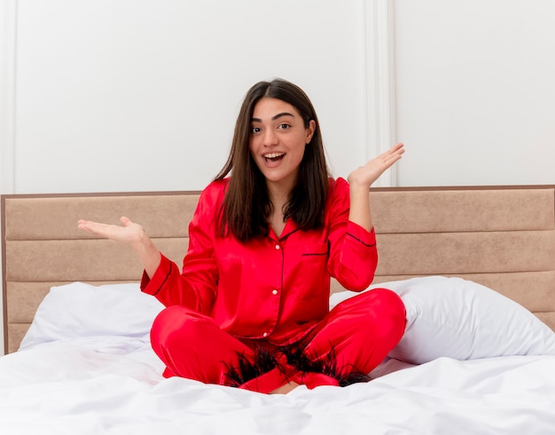 Giovane bella donna in pigiama rosso che si siede sul letto che guarda l'obbiettivo sorridente felice e positivo con le braccia alzate all'interno della camera da letto su sfondo chiaro