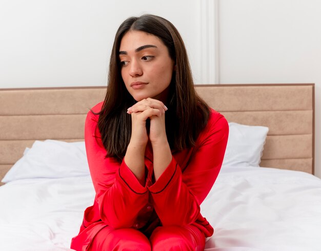 Giovane bella donna in pigiama rosso che si siede sul letto che osserva da parte con l'espressione triste sul viso all'interno della camera da letto su sfondo chiaro