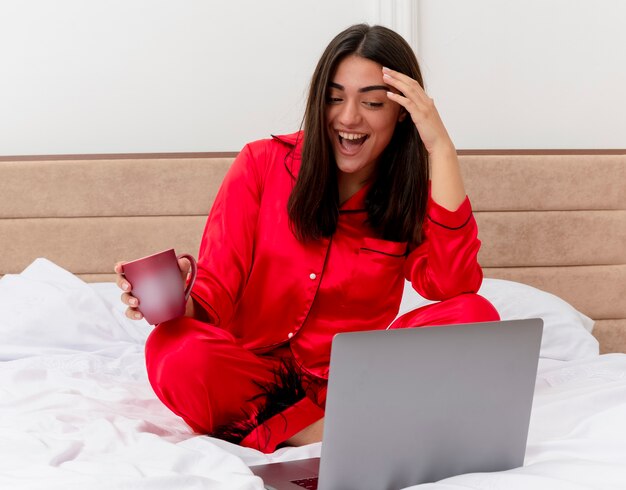 Giovane bella donna in pigiama rosso che si siede sul letto con il computer portatile e la tazza di caffè felice ed emozionato che sorride allegramente nell'interno della camera da letto su sfondo chiaro