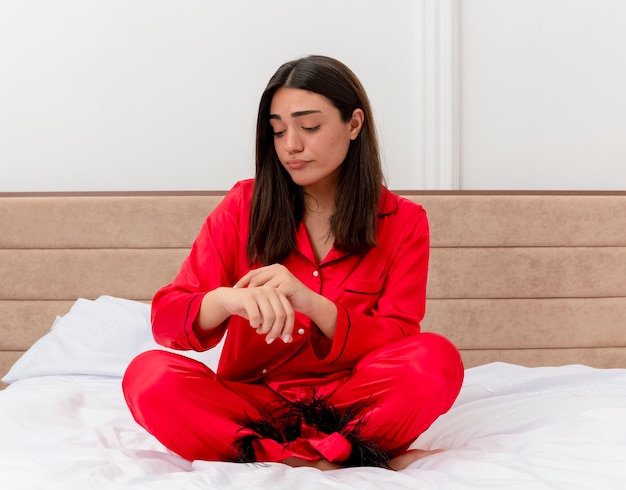 Giovane bella donna in pigiama rosso che si siede sul letto guardando la sua mano che ricorda il tempo dispiaciuto nell'interno camera da letto su sfondo chiaro