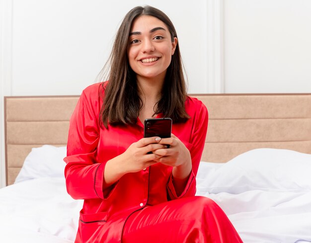 Giovane bella donna in pigiama rosso che si siede sul letto utilizzando smartphone sorridente che guarda l'obbiettivo all'interno della camera da letto su sfondo chiaro