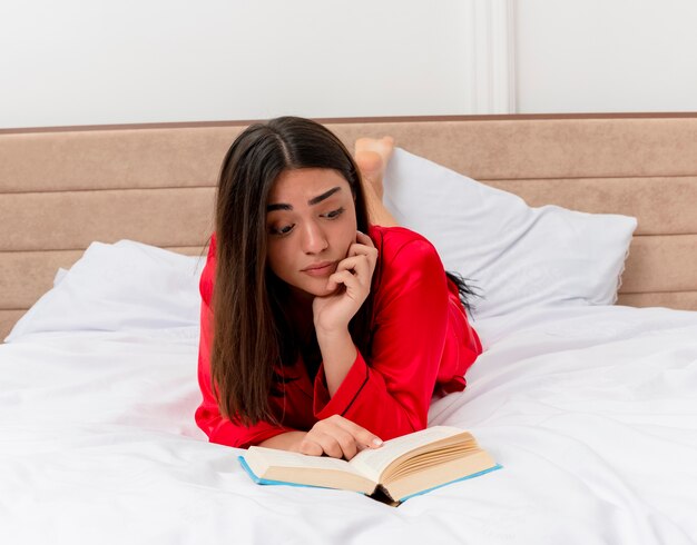 Giovane bella donna in pigiama rosso sdraiato sul letto con un libro che legge con un'espressione triste nell'interno della camera da letto