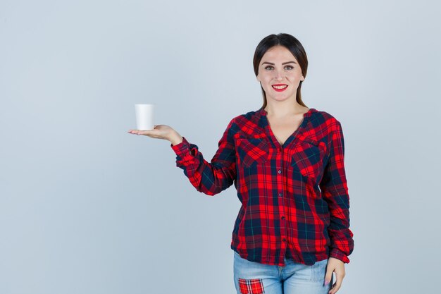 Giovane bella donna in posa mentre tiene in mano un bicchiere di plastica in camicia casual, jeans e sembra allegra, vista frontale.