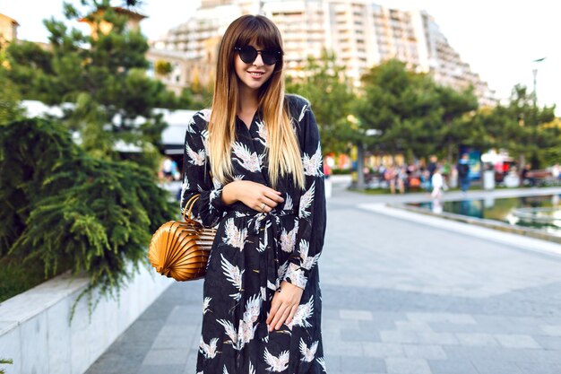 Giovane bella donna in posa sulla strada al centro della città, indossando un elegante abito lungo e una borsa alla moda di paglia vintage, goditi l'atmosfera,