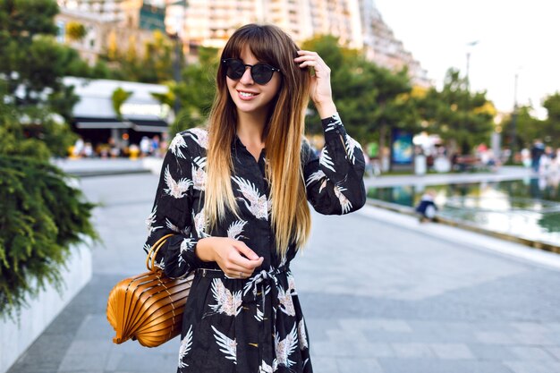 Giovane bella donna in posa sulla strada al centro della città, indossando un elegante abito lungo e una borsa alla moda di paglia vintage, goditi l'atmosfera,