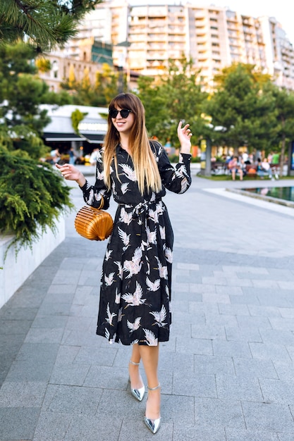 Giovane bella donna in posa sulla strada al centro della città, indossando un elegante abito lungo e una borsa alla moda di paglia vintage, goditi l'atmosfera,