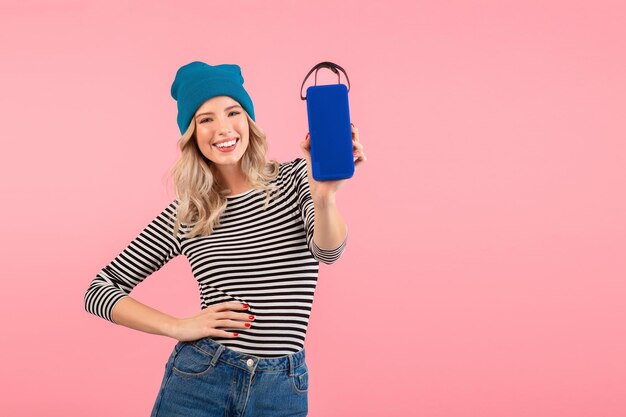 Giovane bella donna in possesso di altoparlante wireless che ascolta musica indossando camicia a righe e cappello blu sorridente felice umore positivo in posa su sfondo rosa isolato