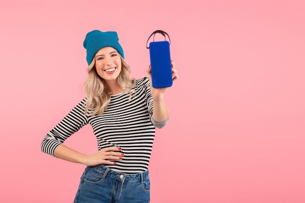 Giovane bella donna in possesso di altoparlante wireless che ascolta musica indossando camicia a righe e cappello blu sorridente felice umore positivo in posa su sfondo rosa isolato