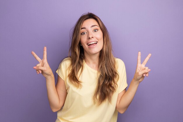 Giovane bella donna in t-shirt beige che guarda l'obbiettivo felice ed eccitata che mostra il v-sign sorridente allegramente in piedi su sfondo viola