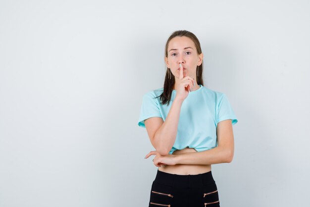 Giovane bella donna in t-shirt, pantaloni che mostrano il gesto del silenzio e sembrano concentrati, vista frontale.