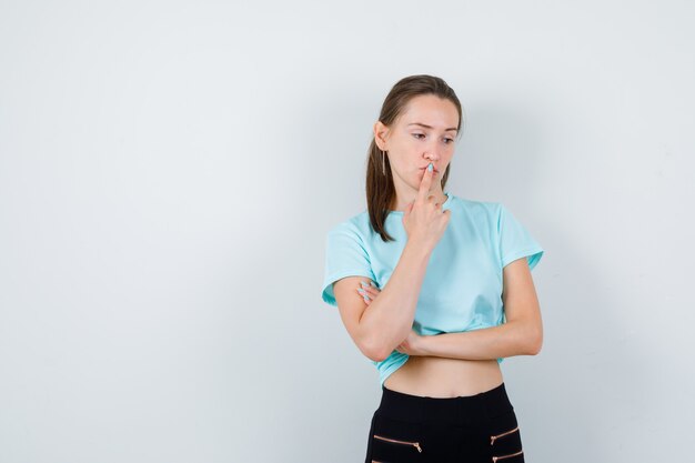 Giovane bella donna in t-shirt, pantaloni con il dito sulla bocca e sguardo pensieroso, vista frontale.