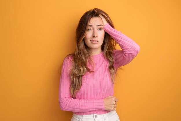 Giovane bella donna in top rosa che guarda la telecamera confusa e molto ansiosa con la mano sulla testa in piedi su sfondo arancione