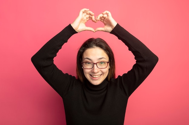 Giovane bella donna in un dolcevita nero e occhiali che fa il gesto del cuore sopra la sua testa sorridendo allegramente in piedi sopra il muro rosa