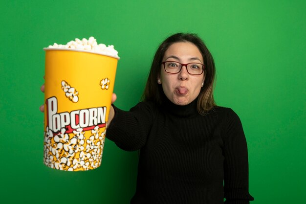 Giovane bella donna in un dolcevita nero e occhiali che mostra secchio con popcorn guardando davanti che attacca fuori la lingua in piedi sopra la parete verde