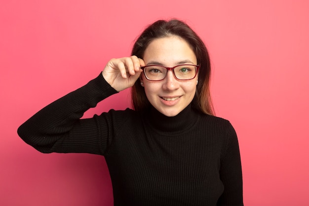 Giovane bella donna in un dolcevita nero e occhiali guardando davanti con il sorriso sul viso in piedi sopra il muro rosa