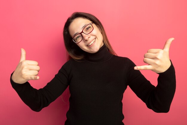 Giovane bella donna in un dolcevita nero e occhiali guardando davanti sorridendo allegramente mostrando chiamami gesto in piedi sopra il muro rosa