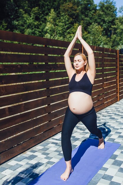 Giovane bella donna incinta che medita con le mani alzate e che cade su un tappetino per lo yoga nella natura sotto il sole