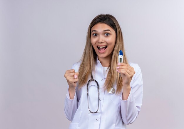 Giovane bella donna medico che indossa camice bianco con lo stetoscopio che tiene il pugno di serraggio termometro digitale uscito e felice