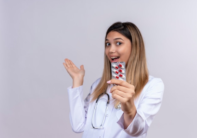 Giovane bella donna medico che indossa camice bianco con lo stetoscopio che tiene la bolla con le pillole che sorride allegramente presentando con il braccio della sua mano