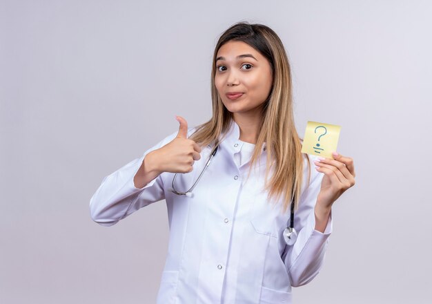 Giovane bella donna medico che indossa camice bianco con lo stetoscopio che tiene la carta di promemoria con il punto interrogativo positivo e felice che mostra i pollici in su