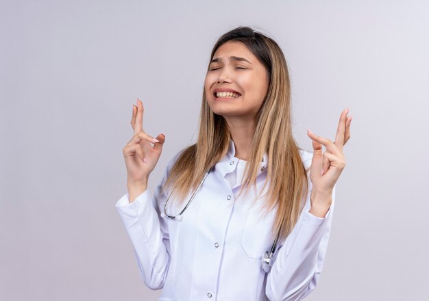 Giovane bella donna medico che indossa camice bianco con lo stetoscopio in piedi con gli occhi chiusi, rendendo desiderabile desiderare dita incrociate con espressione di speranza