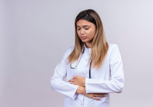 Giovane bella donna medico indossa camice bianco con lo stetoscopio alla ricerca di malessere toccando la sua pancia soffre di dolore