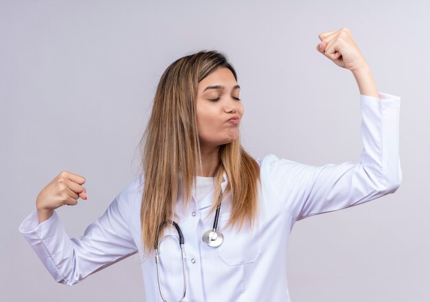 Giovane bella donna medico indossa camice bianco con lo stetoscopio cercando di alzare i pugni fiduciosi in posa come un vincitore