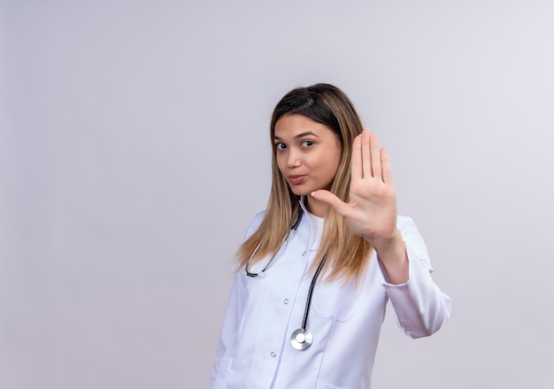 Giovane bella donna medico indossa camice bianco con lo stetoscopio che fa il segnale di stop con la mano aperta con la faccia seria con espressione seria