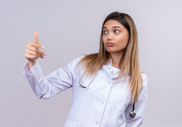 Giovane bella donna medico indossa camice bianco con lo stetoscopio che osserva da parte facendo venire in gesto con la mano