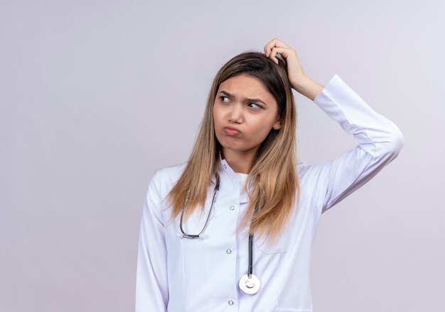 Giovane bella donna medico indossa camice bianco con lo stetoscopio che sembra confuso con il viso accigliato grattandosi la testa avendo dubbi