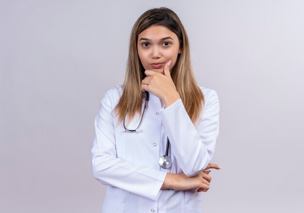 Giovane bella donna medico indossa camice bianco con lo stetoscopio che sembra scontento della mano sul mento in attesa