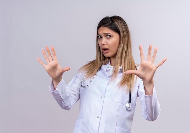 Giovane bella donna medico indossa camice bianco con lo stetoscopio che sembra spaventato facendo gesto di difesa alzando le palme aperte