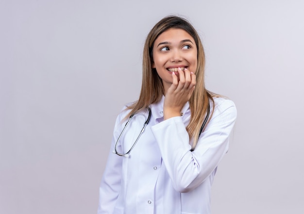 Giovane bella donna medico indossa camice bianco con lo stetoscopio che sembra uscito e felice che morde le unghie in attesa di sorpresa