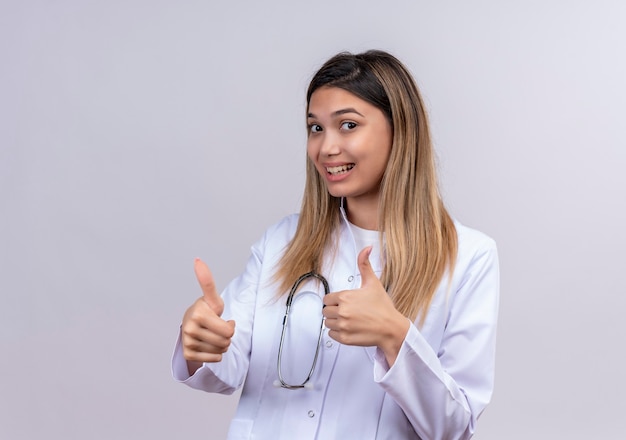 Giovane bella donna medico indossa camice bianco con lo stetoscopio che sorride allegramente mostrando i pollici in su