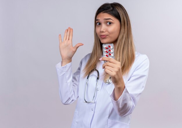 Giovane bella donna medico indossa camice bianco con lo stetoscopio che tiene le pillole della bolla che stanno con la mano aperta che fa il fanale di arresto