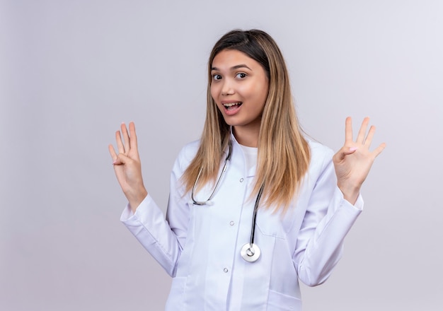 Giovane bella donna medico indossa camice bianco con lo stetoscopio in piedi con le braccia alzate in segno di resa