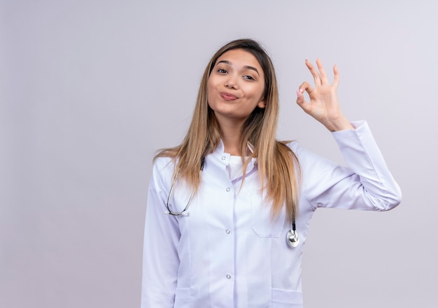 Giovane bella donna medico indossa camice bianco con lo stetoscopio sorridente amichevole facendo segno giusto