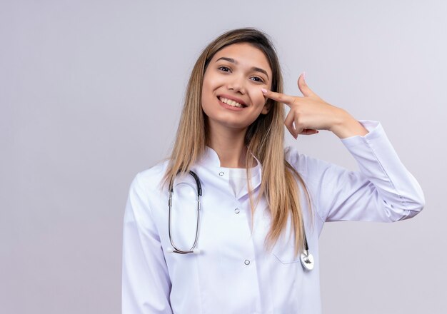 Giovane bella donna medico indossa camice bianco con lo stetoscopio sorridente fiducioso che punta con il dito indice al suo occhio