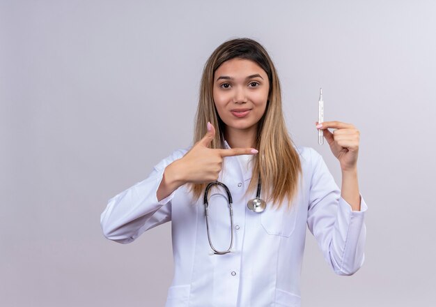Giovane bella donna medico indossa camice bianco con lo stetoscopio tenendo il termometro che punta con il dito indice ad esso guardando fiducioso