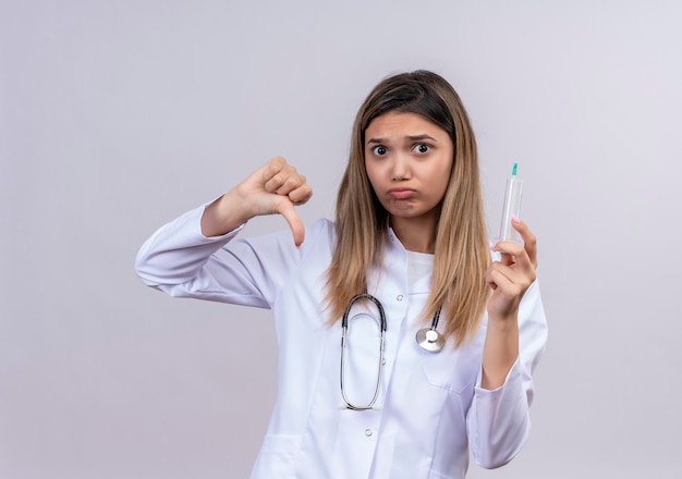 Giovane bella donna medico indossa camice bianco con lo stetoscopio tenendo la siringa guardando con il viso accigliato che mostra i pollici verso il basso