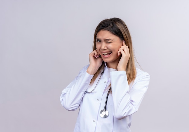 Giovane bella donna medico indossa camice bianco con stetoscopio chiudendo le orecchie con le dita con espressione infastidita per il rumore ad alto volume