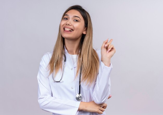 Giovane bella donna medico indossa camice bianco con stetoscopio felice e positivo che punta indietro con il dito indice sorridendo allegramente