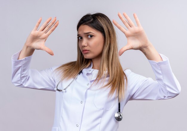 Giovane bella donna medico indossa camice bianco con stetoscopio guardando con faccia seria alzando le mani in segno di resa