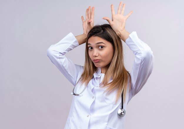Giovane bella donna medico indossa camice bianco con stetoscopio imitando divertenti orecchie da coniglio con le palme sopra la testa