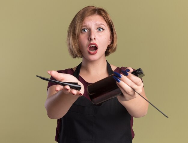 Giovane bella donna parrucchiere in grembiule azienda fermaglio per capelli flacone spray e pettine guardando davanti confuso e deluso in piedi sopra la parete verde