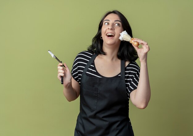 Giovane bella donna parrucchiere in grembiule azienda rasoio e pennello da barba con schiuma sorridente allegramente in piedi sopra la parete chiara