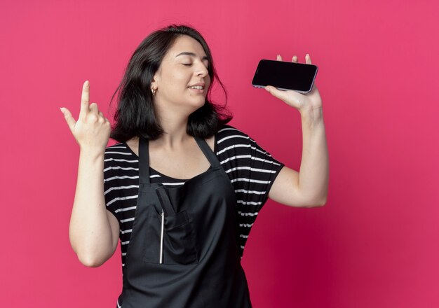 Giovane bella donna parrucchiere in grembiule che tiene smartphone che mostra il dito indice in piedi sopra la parete rosa