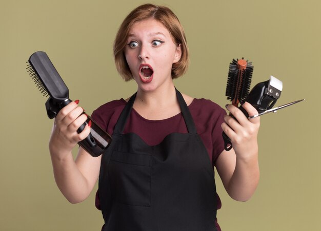 Giovane bella donna parrucchiere in grembiule che tiene trimmer con spazzola per capelli e flacone spray guardando confuso e sorpreso in piedi sopra la parete verde