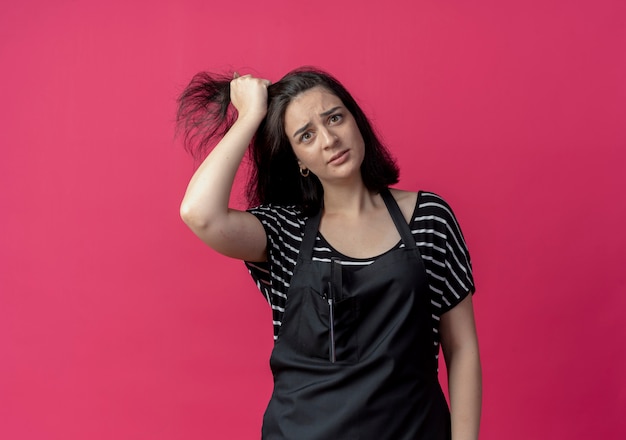 Giovane bella donna parrucchiere in grembiule che tocca i suoi capelli che sembrano confusi sul rosa